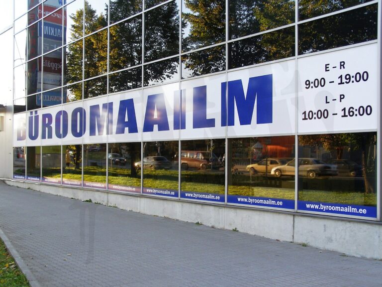 Büroomaailm kaupluse klaasfassaad, millele on paigaldatud suur valge aknakile kirjaga BÜROOMAAILM ja lahtiolekuajad, reklaam akendel.
